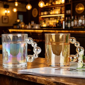 Könnte beinhalten: Zwei transparente Glasbecher, einer mit schillernder Flüssigkeit, der andere bernsteinfarben. Jeder hat einen Griff aus verbundenen Glaskugeln. Sie stehen auf einer Holzoberfläche, mit einer Bar im Hintergrund.
