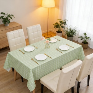 May include: A dining table set with a light green checkered tablecloth, white plates, and gold-colored cutlery. Four cream-colored chairs surround the table. A vase of pink flowers sits in the center. The room is well-lit with natural light.