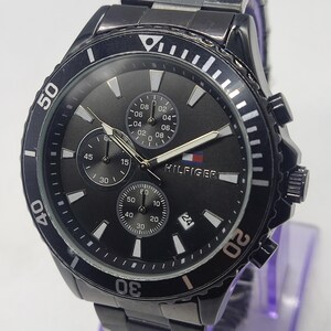 May include: Black Tommy Hilfiger watch with a round face and a black metal band. The watch features a black dial with white markers, three sub-dials, and the Hilfiger logo. The bezel has white markings and the watch has a date display.