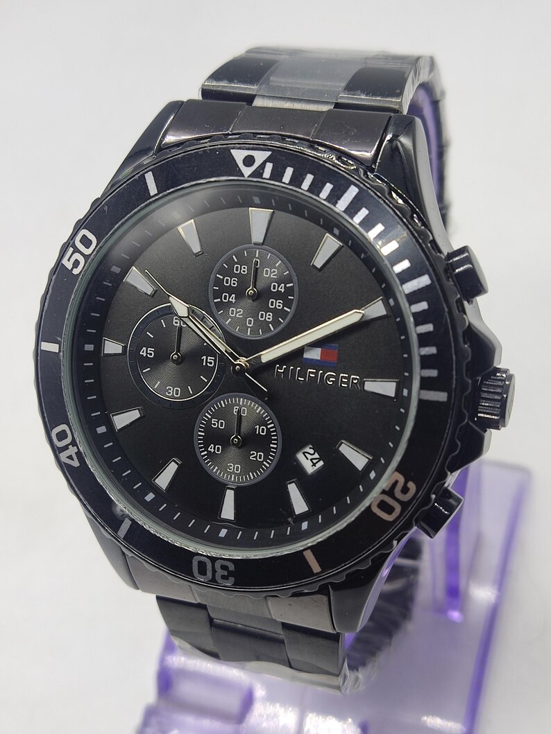 May include: Black Tommy Hilfiger watch with a round face and a black metal band. The watch features a black dial with white markers, three sub-dials, and the Hilfiger logo. The bezel has white markings and the watch has a date display.