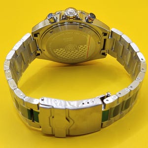 May include: A silver stainless steel wristwatch with a silver band and a yellow background. The watch face is visible, with multiple dials and buttons. The back of the watch has text that reads "Swiss Made Since 1860". The watch band has green accents.