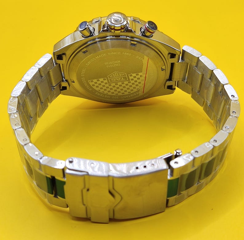 May include: A silver stainless steel wristwatch with a silver band and a yellow background. The watch face is visible, with multiple dials and buttons. The back of the watch has text that reads "Swiss Made Since 1860". The watch band has green accents.
