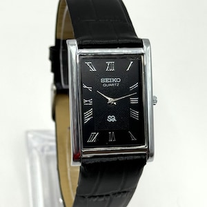 Könnte beinhalten: Rechteckige Seiko Quartz-Uhr mit schwarzem Zifferblatt und silberfarbenem Gehäuse. Die Uhr hat römische Ziffern und ein schwarzes Lederarmband mit Krokodilmuster.