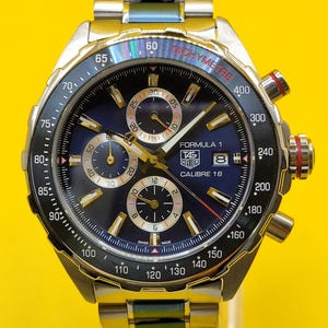 May include: A stainless steel TAG Heuer Formula 1 Calibre 16 watch with a blue dial, gold accents, and a tachymeter bezel. The watch features chronograph subdials and a date display. The band is stainless steel.