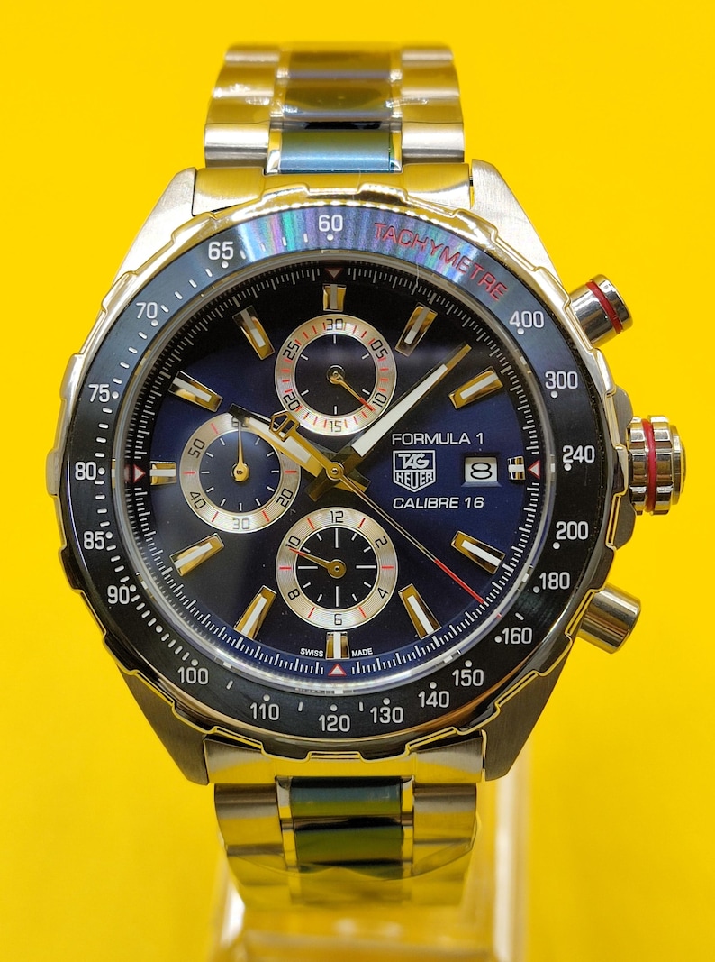 May include: A stainless steel TAG Heuer Formula 1 Calibre 16 watch with a blue dial, gold accents, and a tachymeter bezel. The watch features chronograph subdials and a date display. The band is stainless steel.