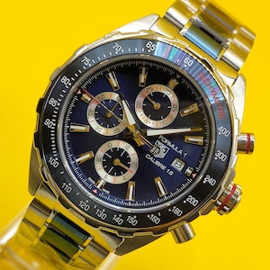 May include: A stainless steel TAG Heuer Formula 1 Calibre 16 watch with a blue dial and a silver-tone stainless steel band. The watch features a tachymeter bezel and three sub-dials. The watch face displays the text "Formula 1" and "Calibre 16".