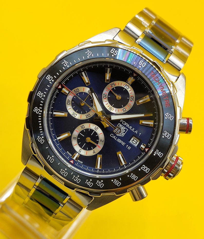 May include: A stainless steel TAG Heuer Formula 1 Calibre 16 watch with a blue dial and a silver-tone stainless steel band. The watch features a tachymeter bezel and three sub-dials. The watch face displays the text "Formula 1" and "Calibre 16".
