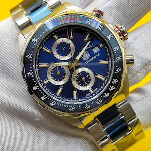 May include: A stainless steel wristwatch with a blue face and gold accents. The watch features multiple sub-dials, a tachymeter bezel, and a date display. The watch band is a combination of silver and blue links.