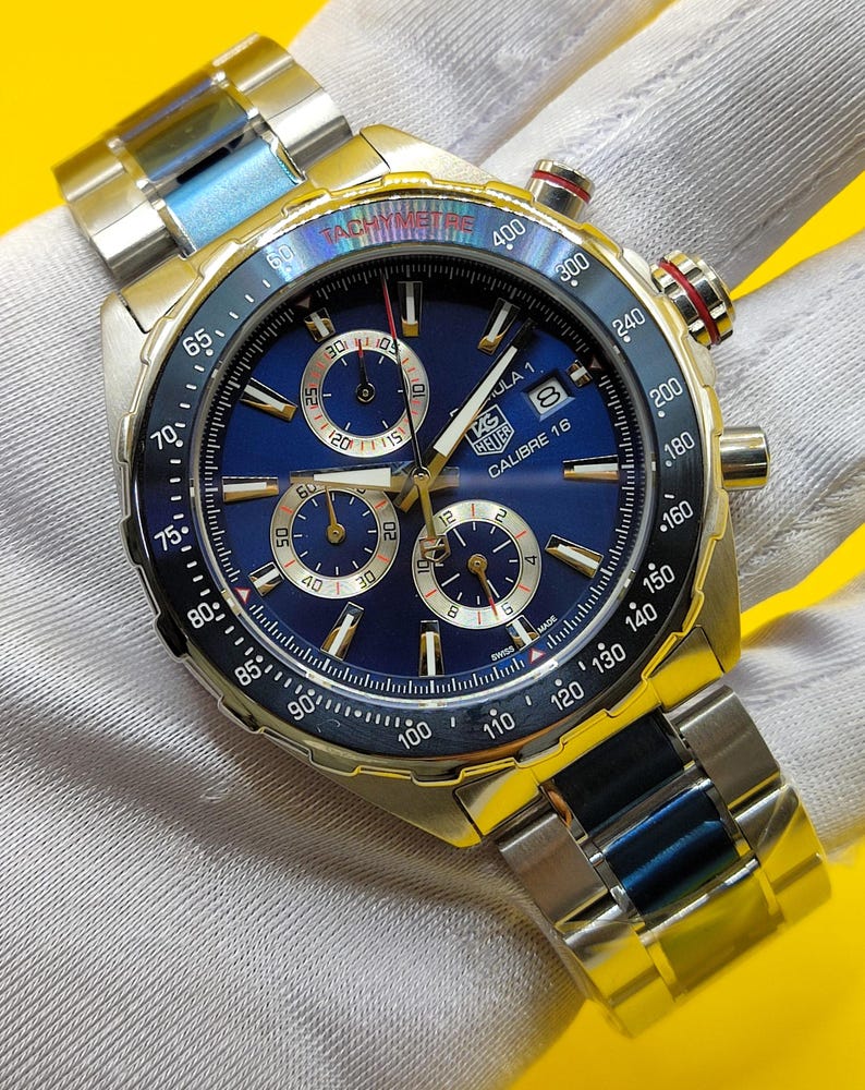May include: A stainless steel wristwatch with a blue face and gold accents. The watch features multiple sub-dials, a tachymeter bezel, and a date display. The watch band is a combination of silver and blue links.