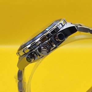 May include: A silver-toned wristwatch with a black face and a stainless steel band. The watch has multiple dials and buttons, and a luminous bezel. The watch is displayed against a bright yellow background.