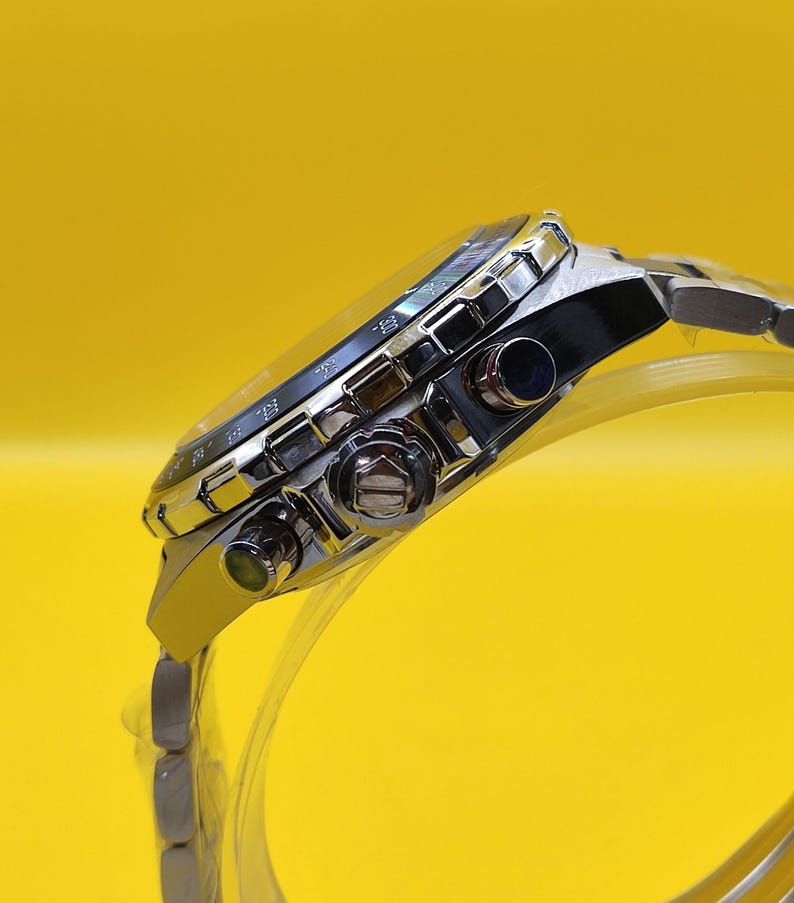 May include: A silver-toned wristwatch with a black face and a stainless steel band. The watch has multiple dials and buttons, and a luminous bezel. The watch is displayed against a bright yellow background.