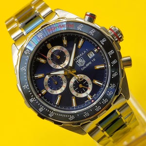 May include: A stainless steel TAG Heuer Formula 1 Calibre 16 watch with a blue dial. The watch features a tachymeter bezel, chronograph subdials, and a metal band. The watch has a red crown and is set against a yellow background.