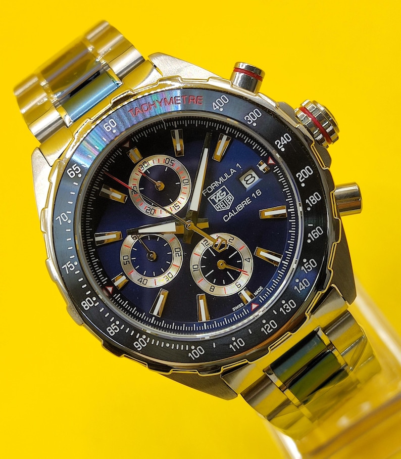 May include: A stainless steel TAG Heuer Formula 1 Calibre 16 watch with a blue dial. The watch features a tachymeter bezel, chronograph subdials, and a metal band. The watch has a red crown and is set against a yellow background.