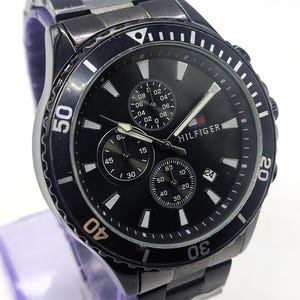 May include: Black stainless steel watch with a black dial. The watch features three sub-dials, white markers, and the brand name "HILFIGER". The watch has a rotating bezel with white numerals and a matching black stainless steel band.