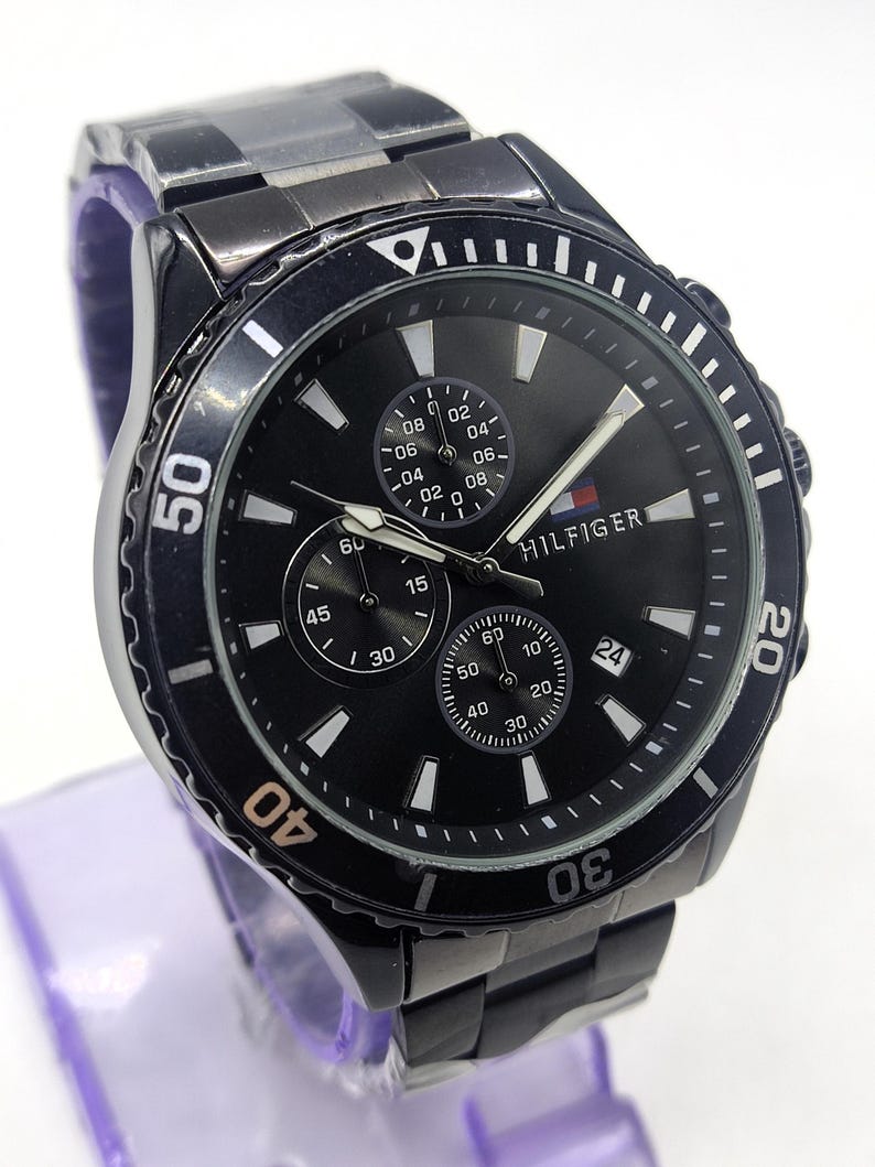 May include: Black stainless steel watch with a black dial. The watch features three sub-dials, white markers, and the brand name "HILFIGER". The watch has a rotating bezel with white numerals and a matching black stainless steel band.