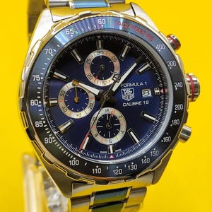 May include: A stainless steel TAG Heuer Formula 1 Calibre 16 watch with a blue dial. The watch features a tachymeter bezel, chronograph subdials, and a date display. The watch band is also stainless steel.