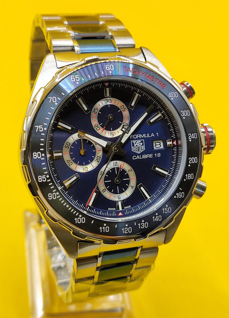 May include: A stainless steel TAG Heuer Formula 1 Calibre 16 watch with a blue dial. The watch features a tachymeter bezel, chronograph subdials, and a date display. The watch band is also stainless steel.