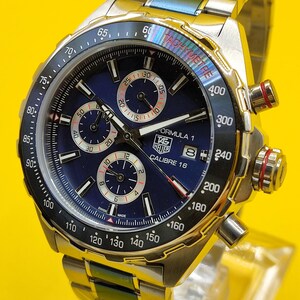 May include: A stainless steel TAG Heuer Formula 1 Calibre 16 watch with a blue dial and chronograph subdials. The watch features a tachymeter bezel and a stainless steel bracelet. The watch is displayed on a clear stand.