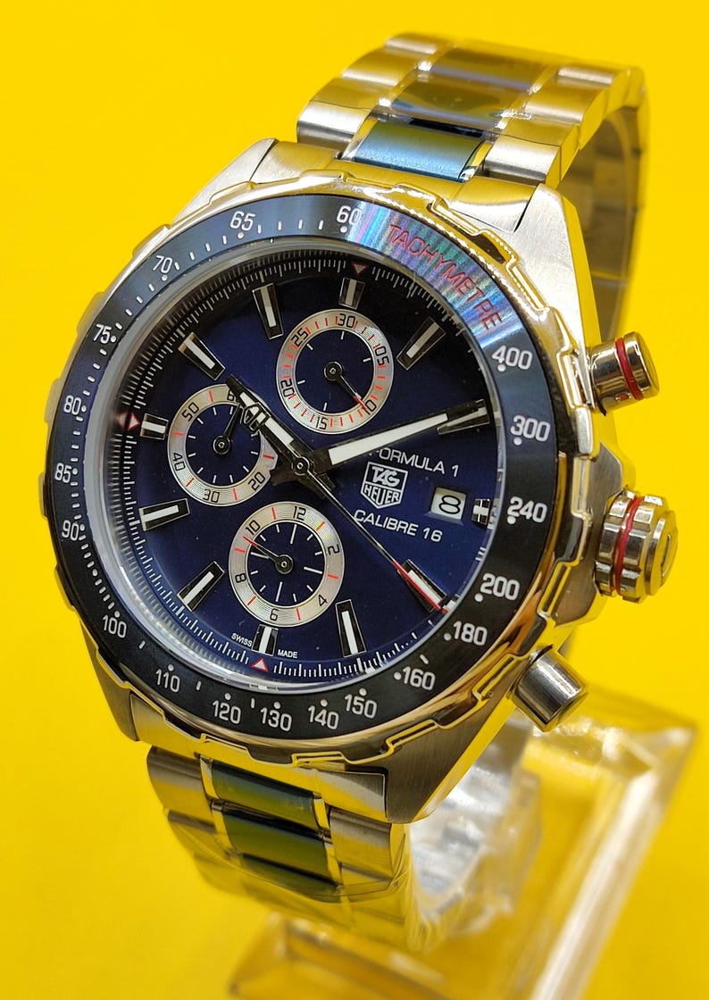 May include: A stainless steel TAG Heuer Formula 1 Calibre 16 watch with a blue dial and chronograph subdials. The watch features a tachymeter bezel and a stainless steel bracelet. The watch is displayed on a clear stand.