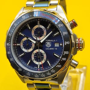 May include: A stainless steel Formula 1 chronograph watch with a blue dial, featuring subdials, a tachymeter scale, and the text "TAG Heuer Calibre 16". The watch has a metal band and red accents on the crown.