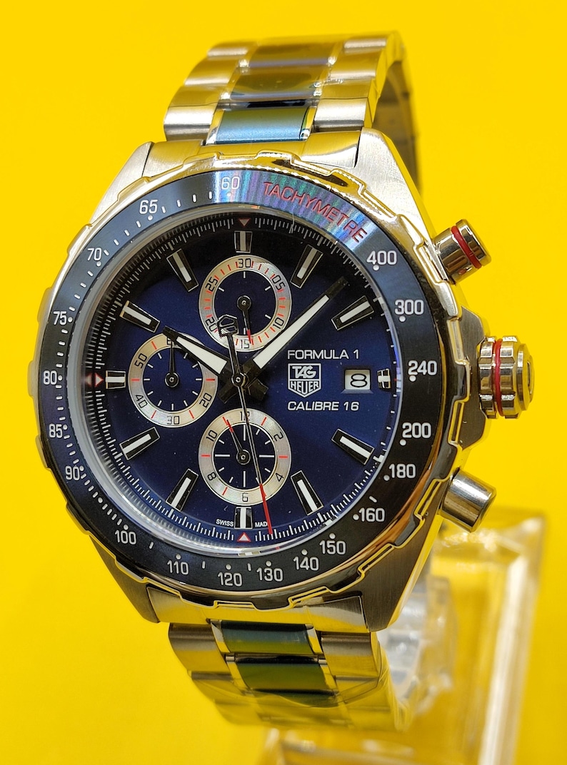 May include: A stainless steel Formula 1 chronograph watch with a blue dial, featuring subdials, a tachymeter scale, and the text "TAG Heuer Calibre 16". The watch has a metal band and red accents on the crown.
