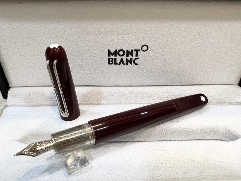 May include: A burgundy Montblanc fountain pen with a silver nib and accents, displayed in an open white box. The pen's cap is off, revealing the nib. The Montblanc logo is visible on the box.