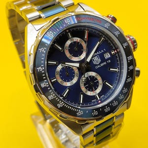 May include: A stainless steel Formula 1 Calibre 16 watch with a blue dial. The watch features a tachymeter bezel and three sub-dials. The watch band is also stainless steel.