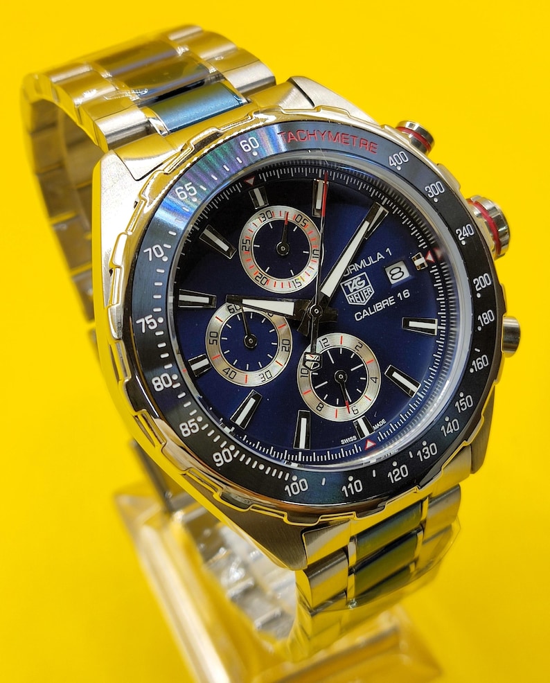 May include: A stainless steel Formula 1 Calibre 16 watch with a blue dial. The watch features a tachymeter bezel and three sub-dials. The watch band is also stainless steel.