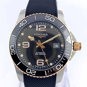 Könnte beinhalten: Eine Longines Automatikuhr mit schwarzem Zifferblatt, goldfarbenen Zeigern und Ziffern sowie einem Datumsfenster auf der Drei-Uhr-Position. Die Uhr verfügt über eine schwarze drehbare Lünette mit goldfarbenem Rand und ein strukturiertes schwarzes Kautschukarmband.