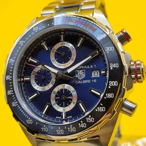 May include: A stainless steel TAG Heuer Formula 1 Calibre 16 watch with a blue dial. The watch features a tachymeter bezel, chronograph subdials, and a stainless steel bracelet. The watch has a Swiss Made label.