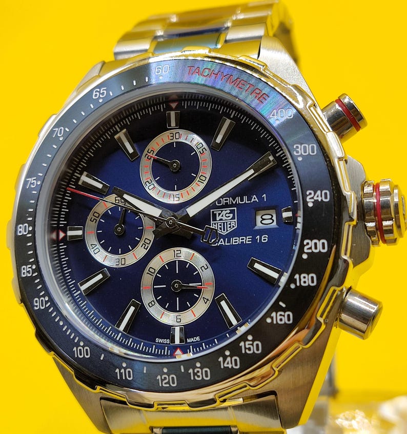 May include: A stainless steel TAG Heuer Formula 1 Calibre 16 watch with a blue dial. The watch features a tachymeter bezel, chronograph subdials, and a stainless steel bracelet. The watch has a Swiss Made label.