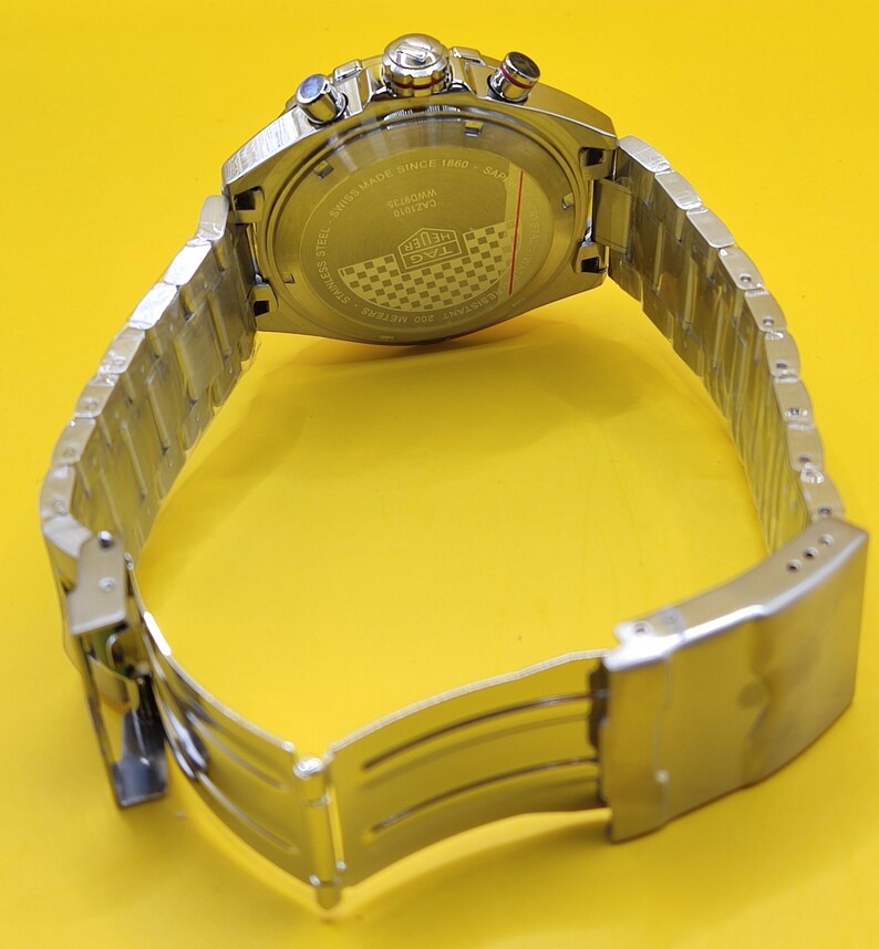 May include: A stainless steel wristwatch with a silver band and a silver watch face. The watch has a round face with multiple dials and buttons. The back of the watch has text that reads "Swiss Made Since 1860".