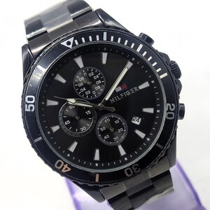 May include: A black Tommy Hilfiger watch with a black dial, featuring multiple sub-dials and a date display. The watch has a black bezel with white markings and a stainless steel band. The watch is displayed on a purple surface.