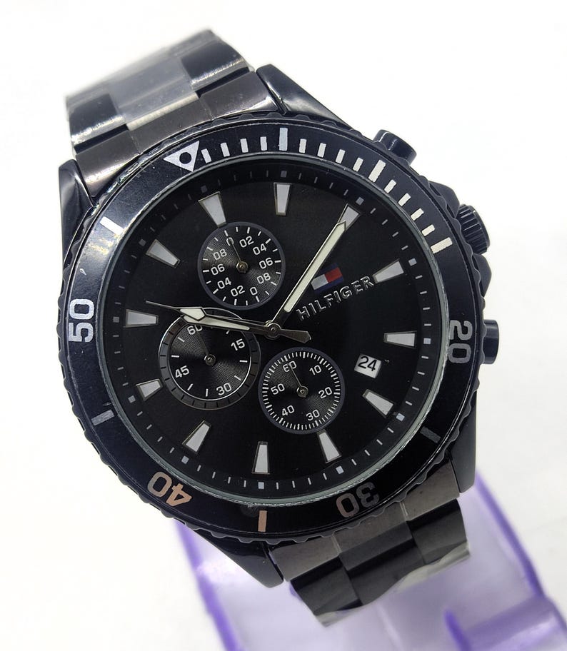 May include: A black Tommy Hilfiger watch with a black dial, featuring multiple sub-dials and a date display. The watch has a black bezel with white markings and a stainless steel band. The watch is displayed on a purple surface.