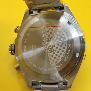 May include: A stainless steel TAG Heuer watch back with the text "SAPPHIRE CRYSTAL - WATER RESISTANT 200 METERS". The watch is marked "SWISS MADE SINCE 1860". The watch has a checkered pattern.