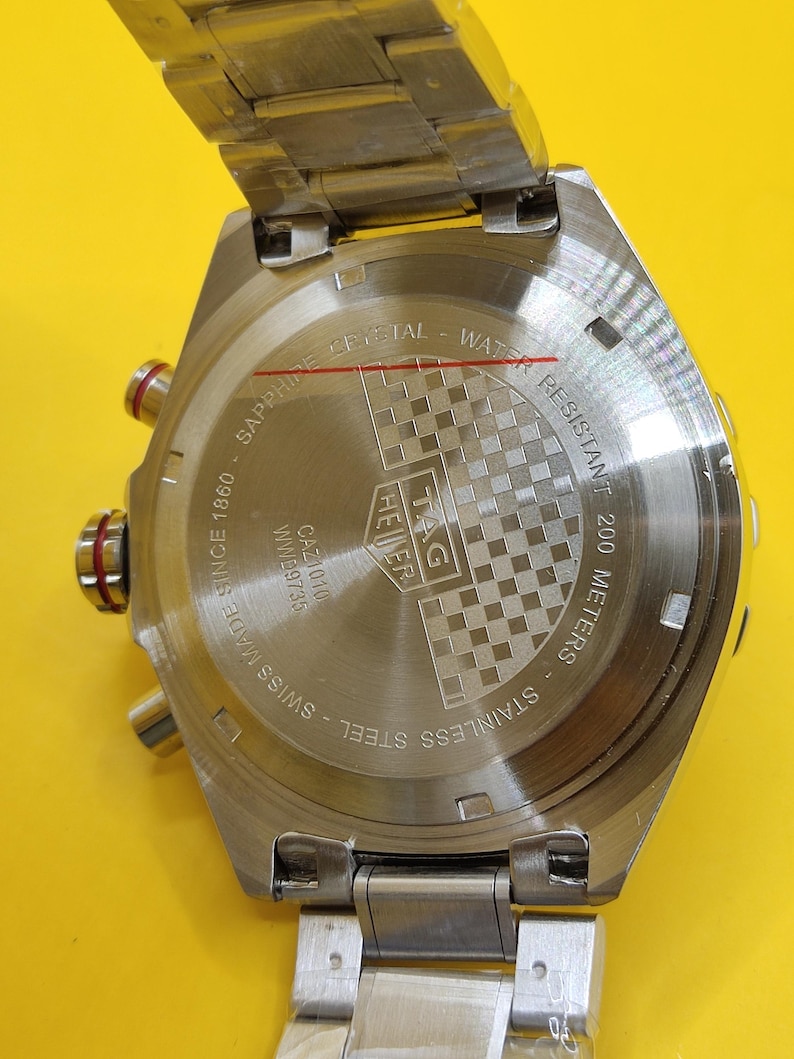 May include: A stainless steel TAG Heuer watch back with the text "SAPPHIRE CRYSTAL - WATER RESISTANT 200 METERS". The watch is marked "SWISS MADE SINCE 1860". The watch has a checkered pattern.