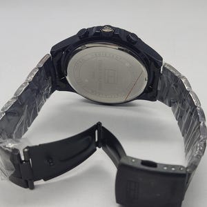May include: A black stainless steel watch with a matching band. The watch face is visible, with the words "Water Resistant" and "Tommy Hilfiger" printed on it. The band is open, showing the clasp.