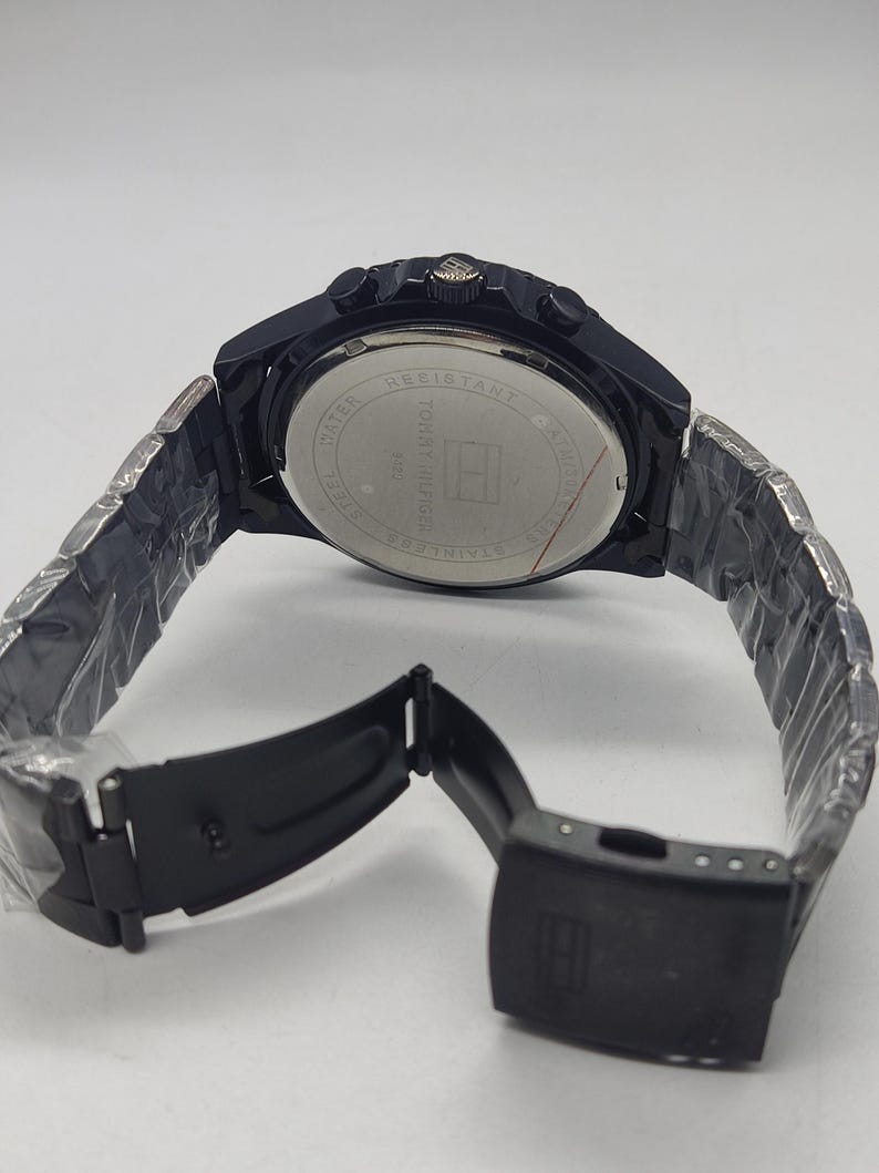 May include: A black stainless steel watch with a matching band. The watch face is visible, with the words "Water Resistant" and "Tommy Hilfiger" printed on it. The band is open, showing the clasp.
