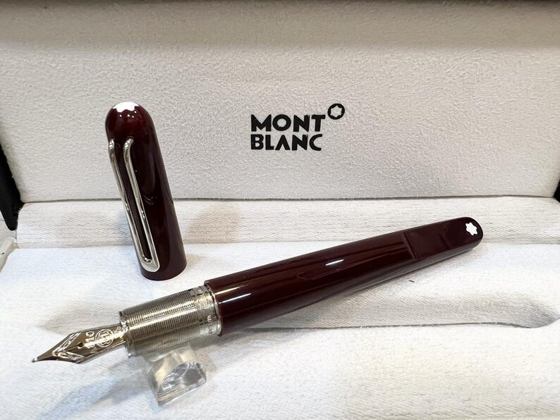 May include: A burgundy Montblanc fountain pen with a silver nib and accents, displayed in an open white box. The pen's cap is off, revealing the nib. The Montblanc logo is visible on the box.