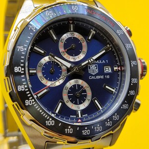 May include: A stainless steel TAG Heuer Formula 1 Calibre 16 watch with a blue dial. The watch features a tachymeter bezel, three sub-dials, and a date window. The watch has a silver band and is set against a yellow background.