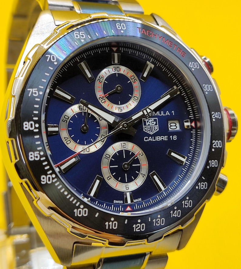 May include: A stainless steel TAG Heuer Formula 1 Calibre 16 watch with a blue dial. The watch features a tachymeter bezel, three sub-dials, and a date window. The watch has a silver band and is set against a yellow background.