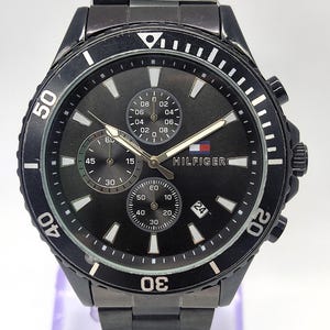 May include: Black Tommy Hilfiger watch with a black dial, featuring three sub-dials and a date display at the 3 o'clock position. The watch has a black bezel with white markers and a black metal band. The Hilfiger logo is visible on the dial.