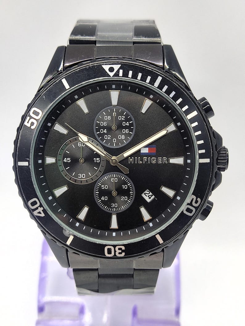 May include: Black Tommy Hilfiger watch with a black dial, featuring three sub-dials and a date display at the 3 o'clock position. The watch has a black bezel with white markers and a black metal band. The Hilfiger logo is visible on the dial.