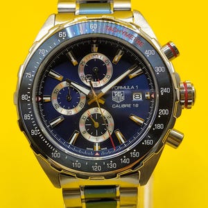 May include: A stainless steel TAG Heuer Formula 1 Calibre 16 watch with a blue dial. The watch features a tachymeter bezel and three sub-dials. The watch band is also stainless steel.