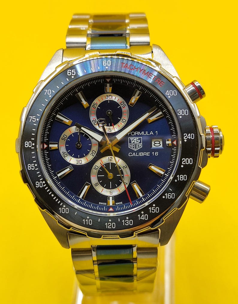 May include: A stainless steel TAG Heuer Formula 1 Calibre 16 watch with a blue dial. The watch features a tachymeter bezel and three sub-dials. The watch band is also stainless steel.