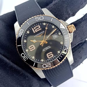 Könnte beinhalten: Eine Longines Automatikuhr mit schwarzem Zifferblatt, goldfarbenen Zeigern und Ziffern sowie einem Datumsfenster auf der Drei-Uhr-Position. Die Uhr verfügt über eine drehbare schwarze Keramiklünette mit goldfarbenen Akzenten, ein Edelstahlgehäuse und ein schwarzes, strukturiertes Kautschukarmband.