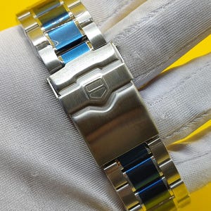 May include: Close-up of a stainless steel watch band with blue accents. The band features a secure clasp and a polished finish. The watch band is designed for durability and style.