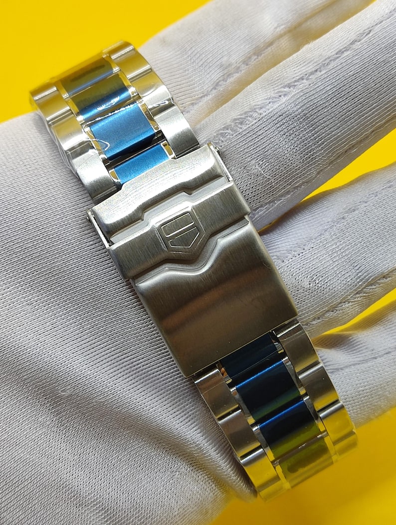 May include: Close-up of a stainless steel watch band with blue accents. The band features a secure clasp and a polished finish. The watch band is designed for durability and style.