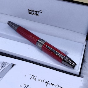 Könnte beinhalten: Ein roter Montblanc-Stift in einer schwarz-weißen Box. Der Stift hat einen strukturierten roten Schaft, silberne Akzente und eine schwarze Spitze. Die Box ist geöffnet und zeigt den Stift und den Schriftzug "MONTBLANC". Der Text "The art of writing" ist sichtbar.