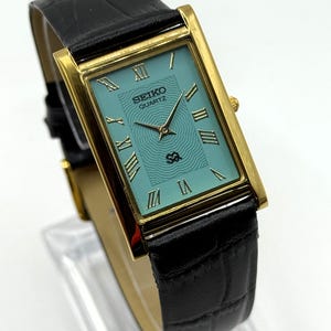 Könnte beinhalten: Eine rechteckige, goldfarbene Seiko Quartz-Uhr mit türkisblauem Zifferblatt und schwarzem Lederarmband. Das Zifferblatt hat römische Ziffern und das Seiko-Logo. Das Armband ist mit einem Krokodilmuster versehen.